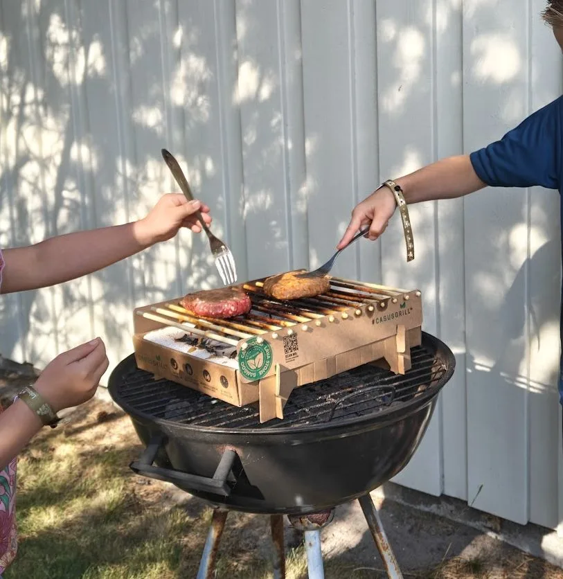 Kinderen draaien hamburgers om op de CasusGrill, met het #CASUSGRILL-logo zichtbaar op de kartonnen barbecue